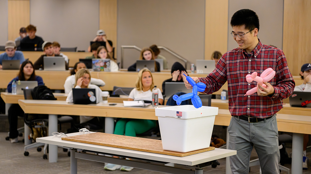 A man holds two balloon animals over a Styrofoam cooler at the front of a tiered classroom.