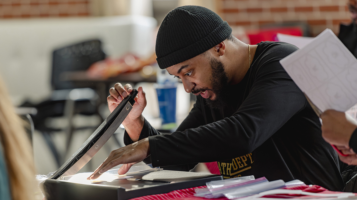 A young man wearing a black cap uses a paper cutter.