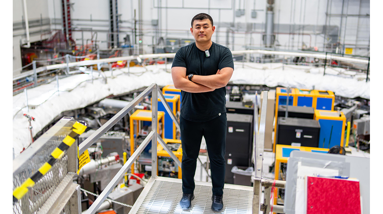 A young man stands on an elevated platform overlooking a room full of high-tech equipment.