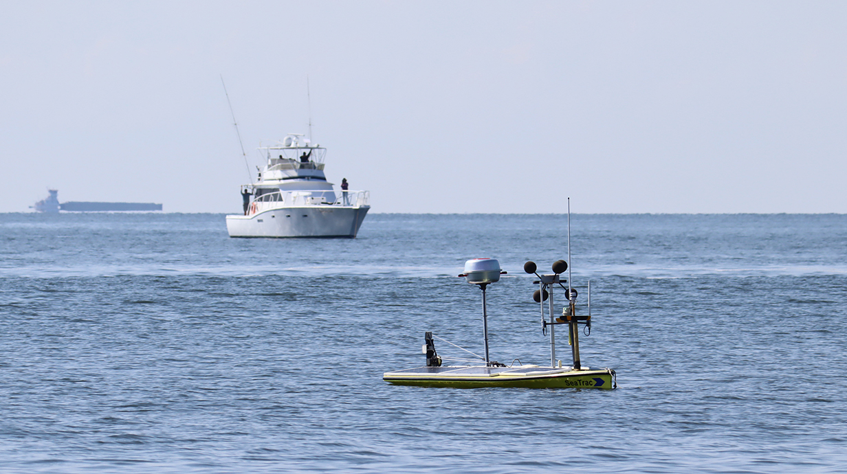 A large boat with people standing on deck follows a smaller, unmanned vehicle on the surface of the ocean.