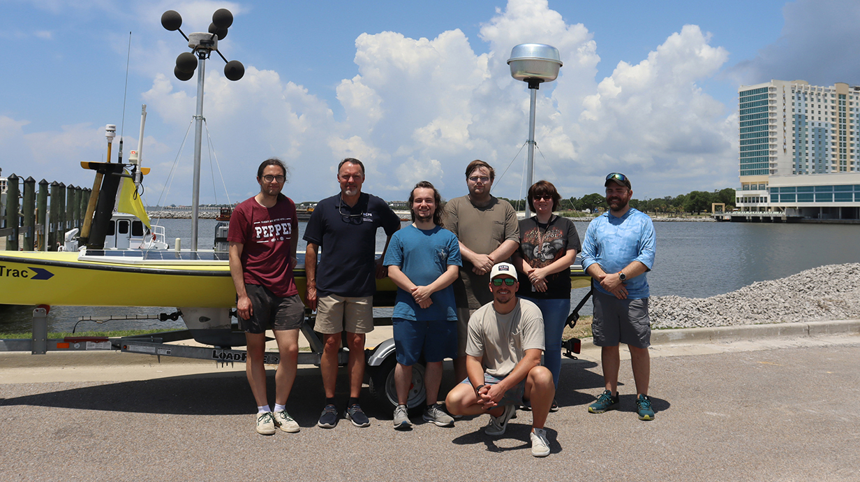 A group of people stands in front of a small watercraft on a coastline.