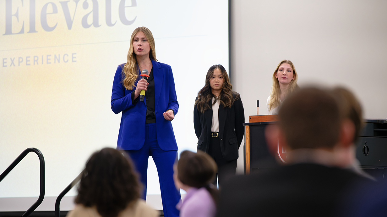 A young woman standing in front of a projection screen on a stage speaks into a microphone as a crowd listens.