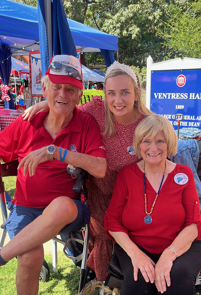 A man and two women, all wearing Ole Miss gear, sit underneath a tailgating tent in front of a sign for Ventress Hall.