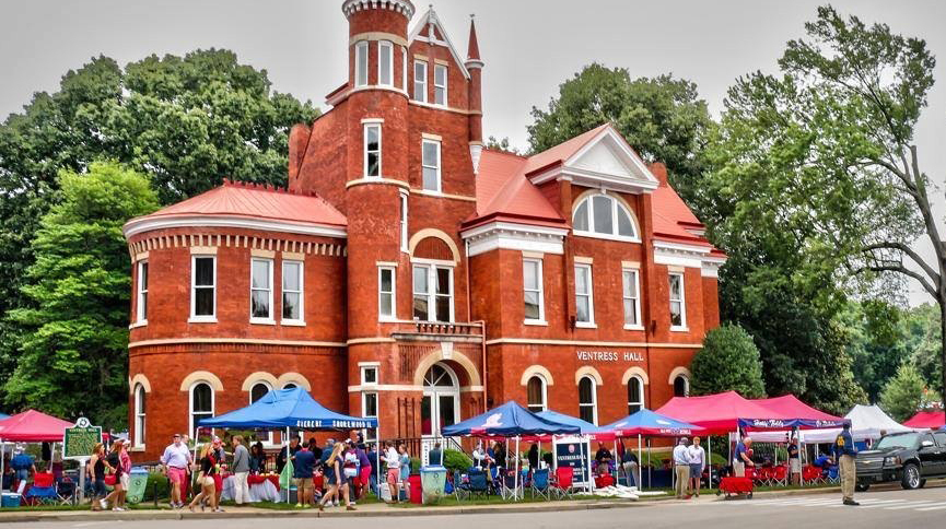 Red and blue tents surround a gothic red-brick building on a tree-lined street.