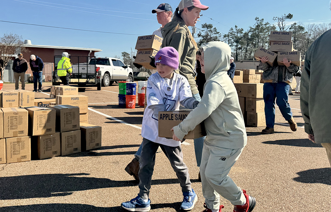 Two young boys carry a box marked 'Apple Sauce' across a parking lot, surrounded by other people doing the same.