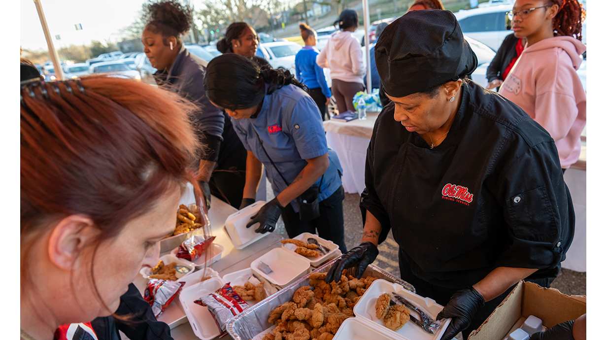 A woman wearing a black chef's jacket helps hand out chicken tenders in foam clamshell containers underneath a tent.