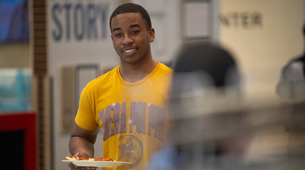 A young man wearing a gold T-shirt carries a tray of food in a dining hall.