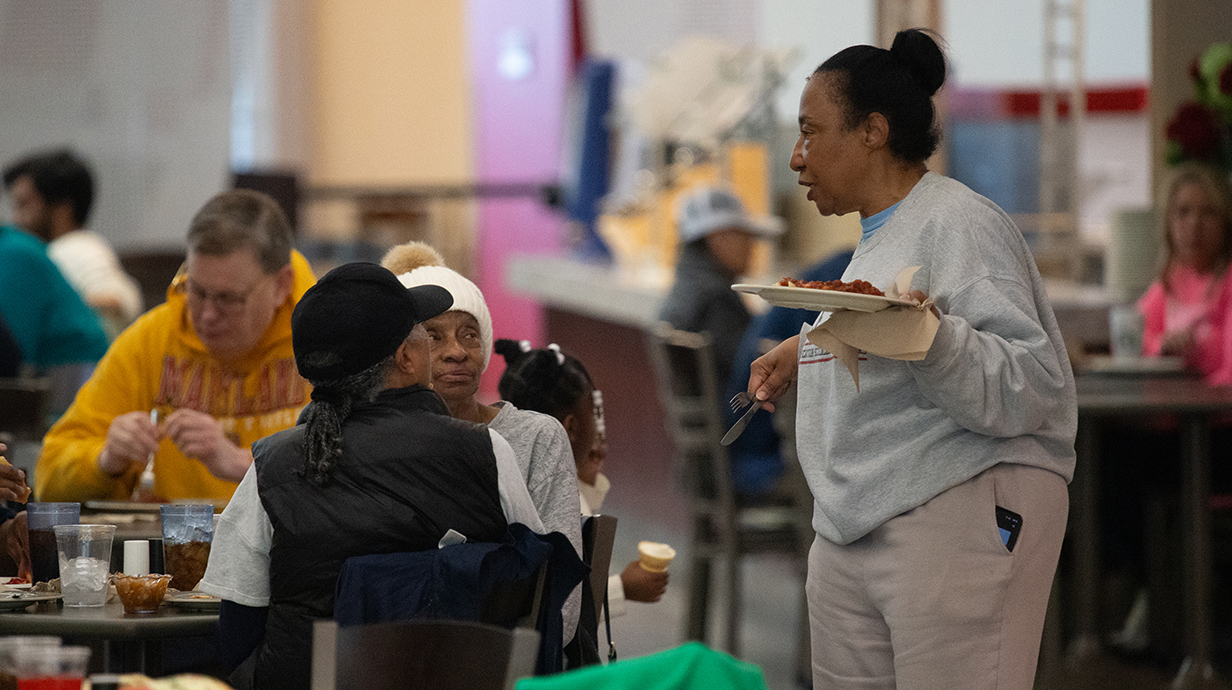 A woman dressed in a sweatsuit and holding a tray of food talks to two women seated at a table.