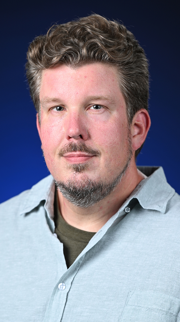 Headshot of a man wearing a light blue buttoned shirt over a brown T-shirt.