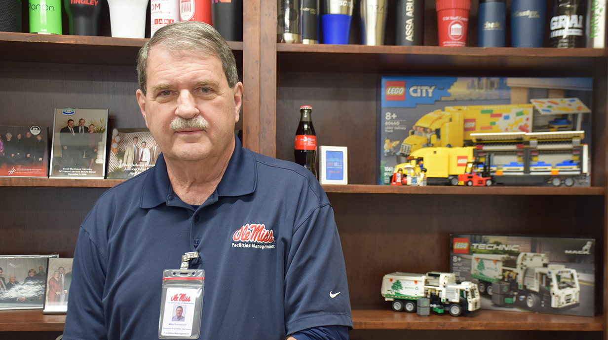 A man wearing a blue polo shirt stands in front of bookcases loaded with books and mementos.