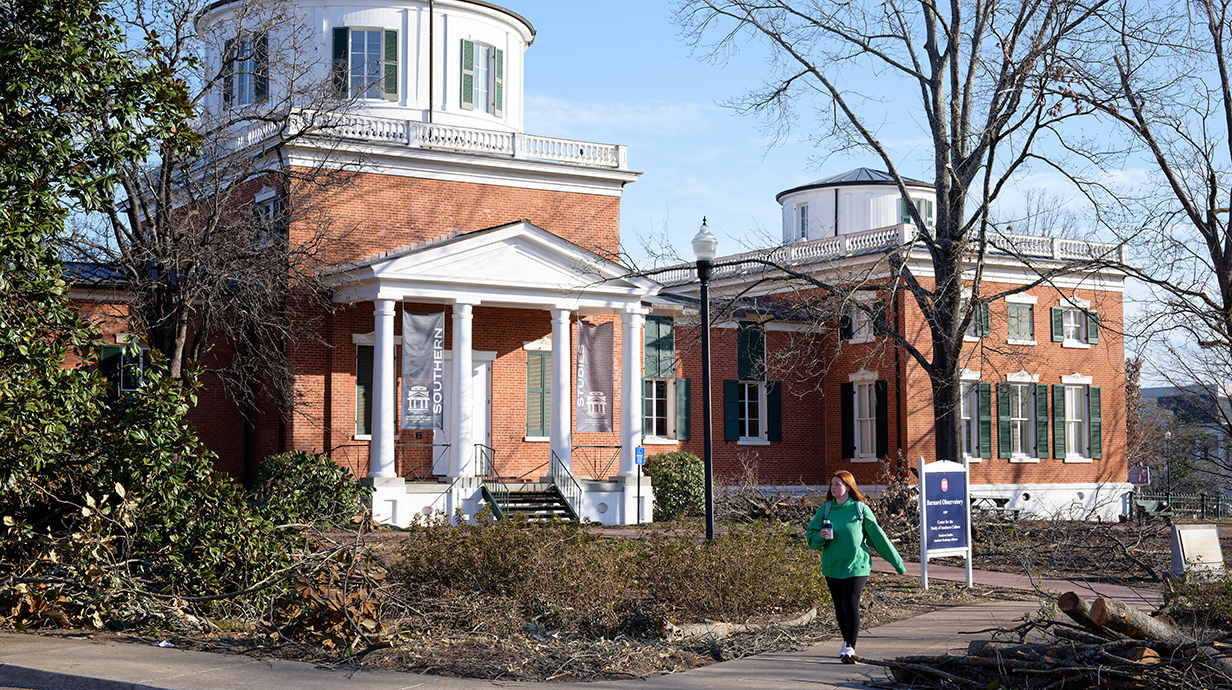 A woman walks past piles of tree debris in front of a large brick building with a white porch and upper floor.