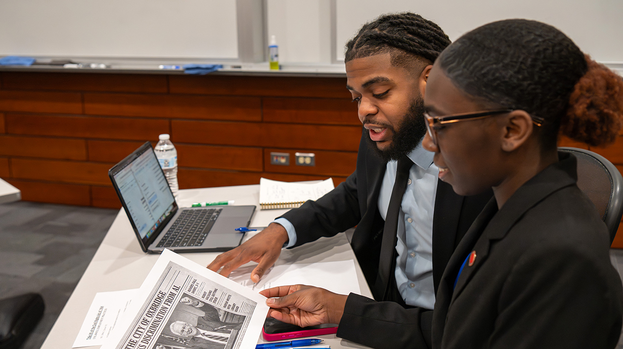 A young man and a young woman, both dressed in business attire, look over paperwork while seated at a table.
