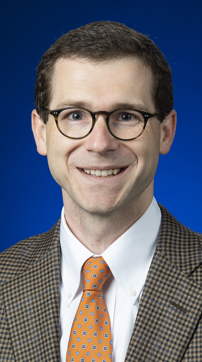 Headshot of a man wearing glasses and a brown suit.