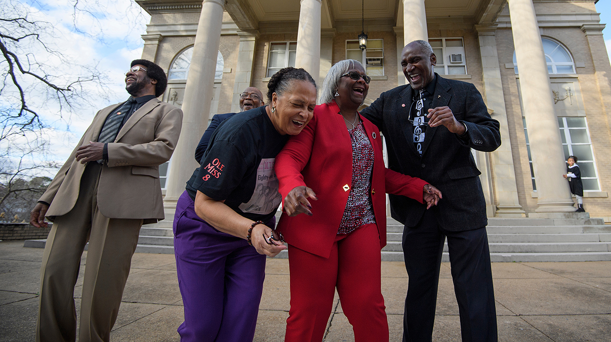 Two women and two men share a hearty laugh outside a large tan stone building.