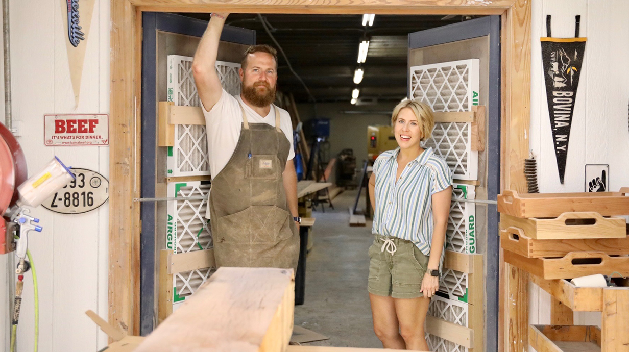 A bearded man wearing tan overalls and a woman wearing a white T-shirt stand in the entrance to a woodshop.