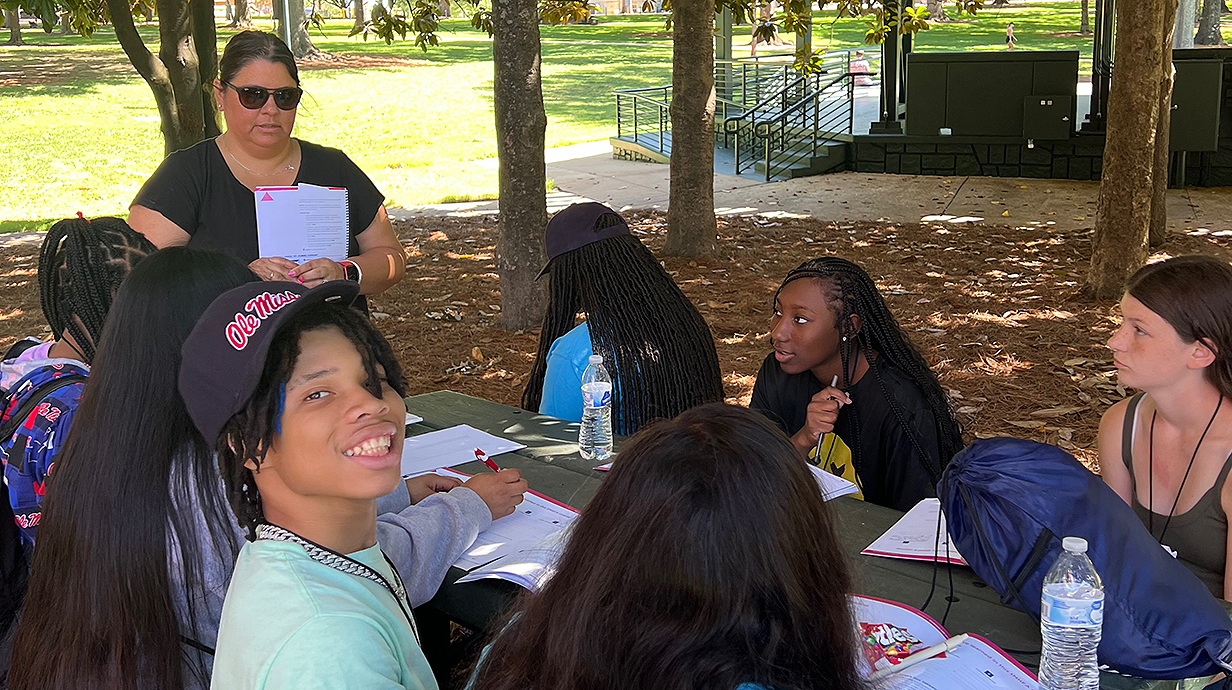 A woman standing at one end of an outdoor picnic table holds up paperwork for a group of young people seated around it.