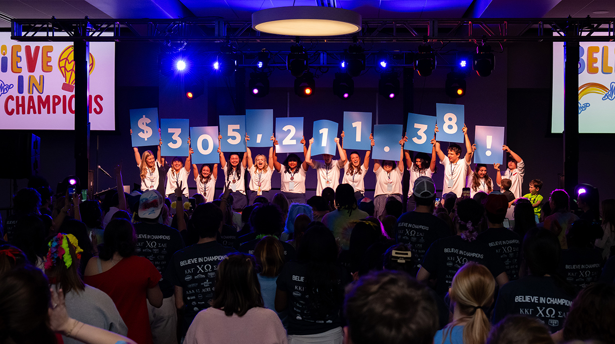 A group of young people standing on a stage in a crowded ballroom hold up cards spelling out '$305,211.38 .'