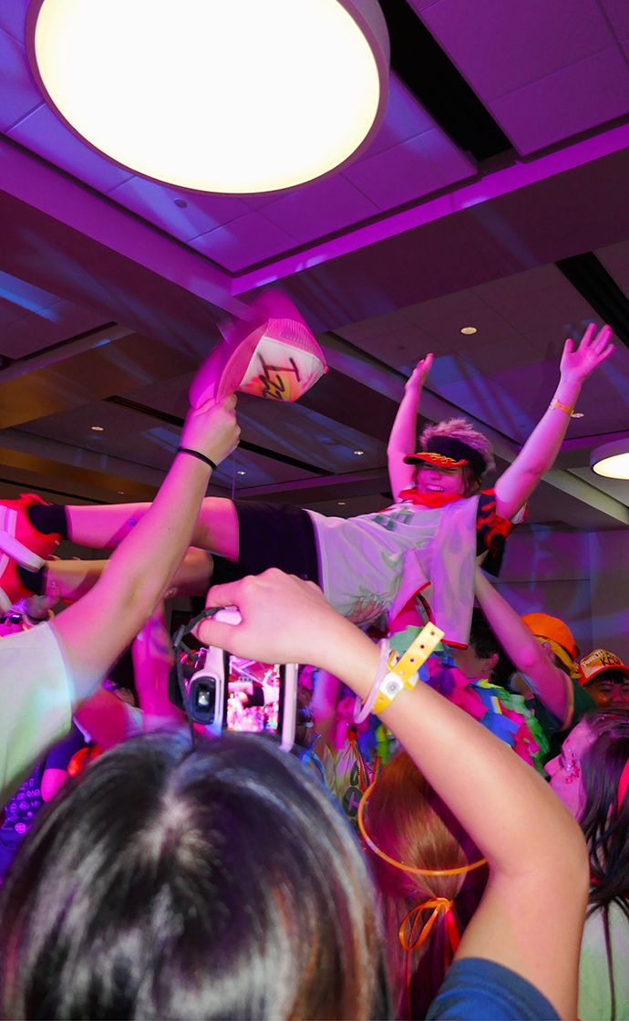 A group of people on a crowded dance floor raise a young woman wearing a spiky wig and sunglasses over their heads.