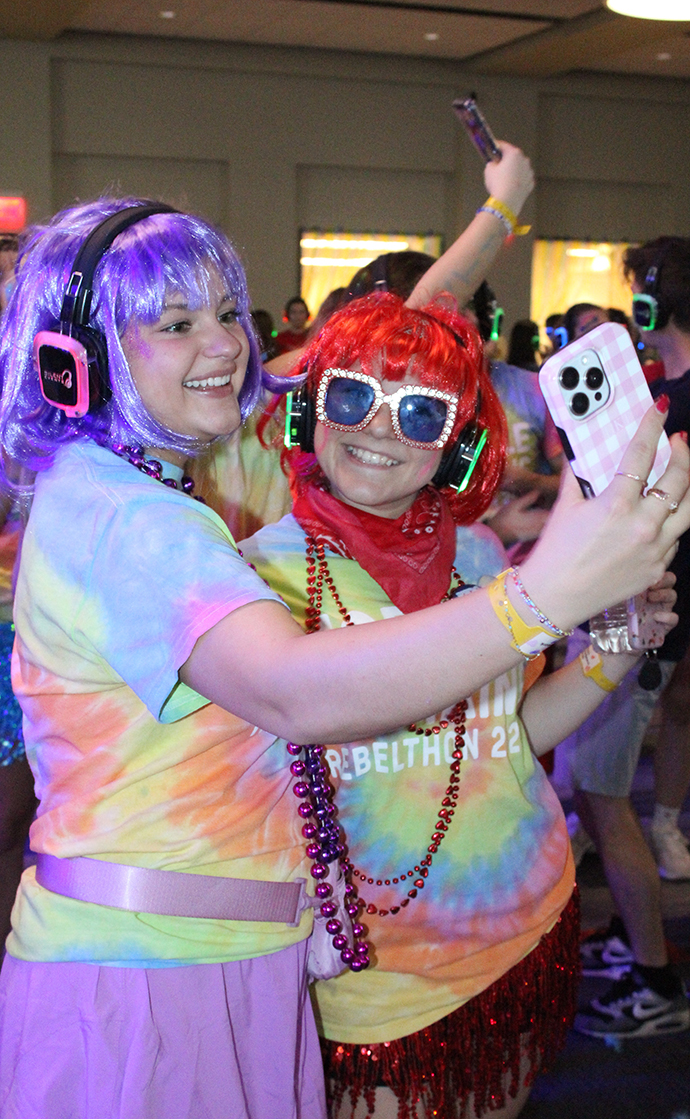 Two young women wearing colorful wigs and costumers take a selfie on a dance floor.