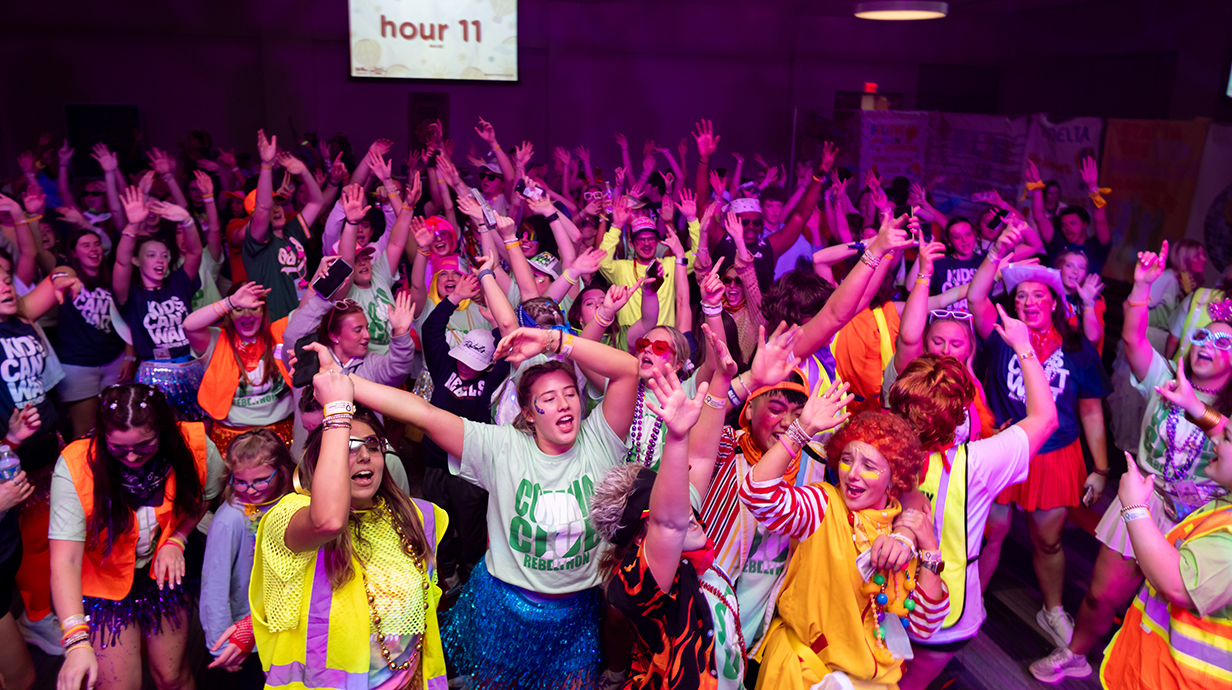 Hundreds of young people dance and sing on a dance floor bathed in colored lights.