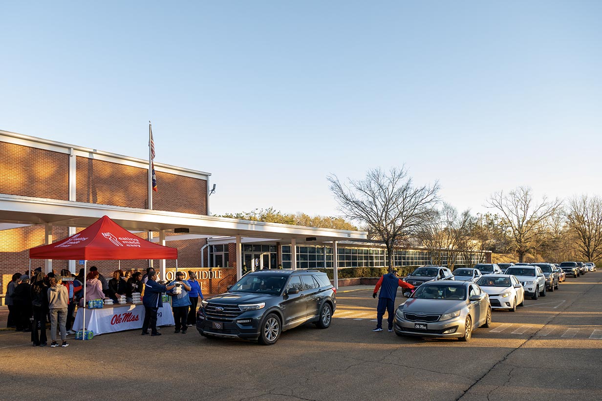 A long line of cars queuing at dusk in front of Oxford Middle School, with staff visible in the distance distributing meal boxes.