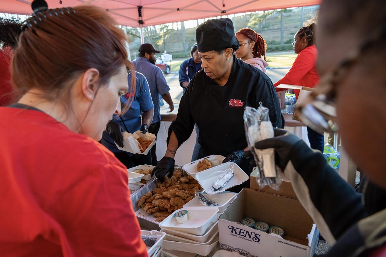 Sous chef Josephine Toliver wearing a mask and apron, focused on packing fried chicken and sides into Styrofoam containers inside a commercial kitchen.