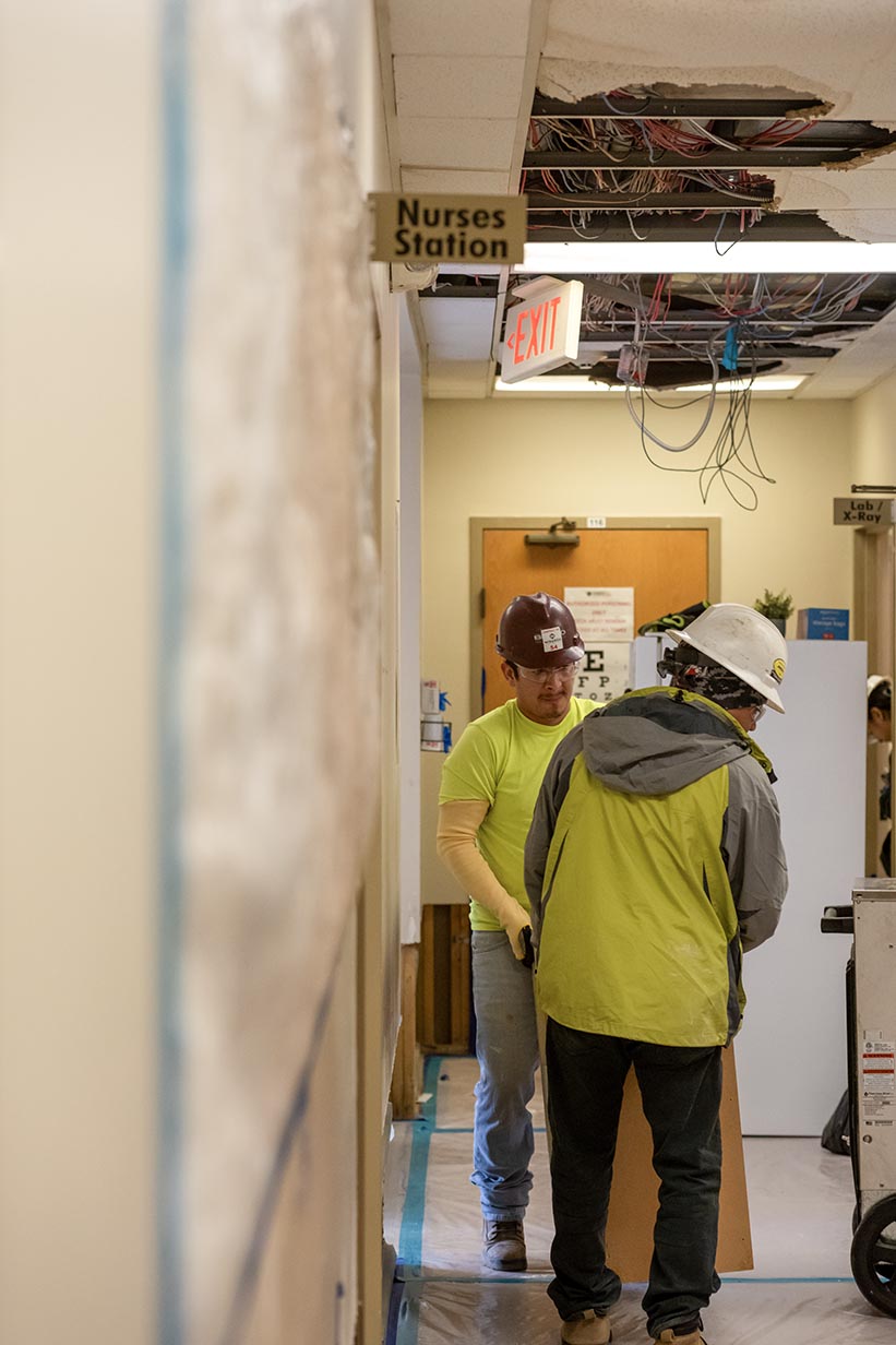 Workers in high-visibility vests moving metal shelving and office furniture out of a clinical room with X-ray equipment.