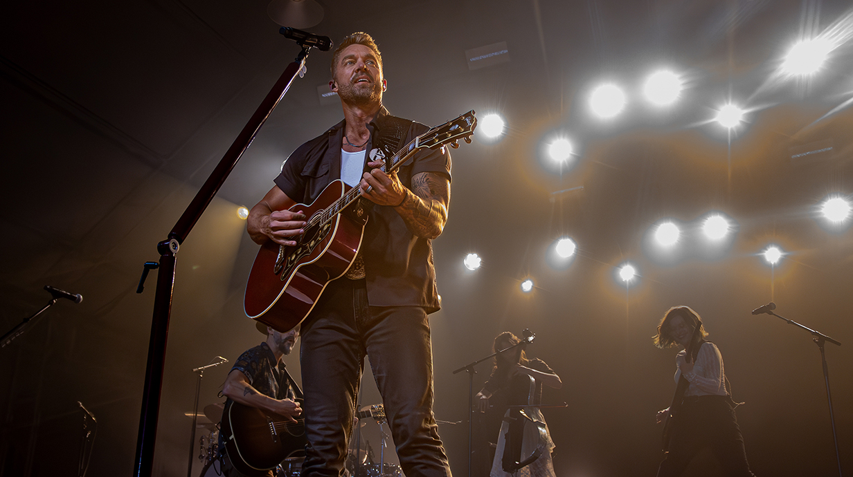 A man plays guitar on a stage with lights and a backing band behind him.