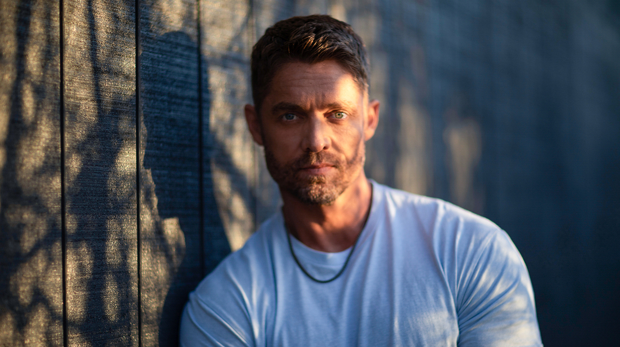 A man wearing a white T-shirt leans against a wall.