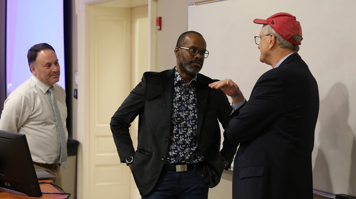 Three men talk while standing in front of a whiteboard in a classroom.
