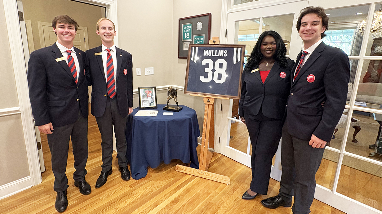 Three young men and a young woman, all wearing navy blue blazers, stand with a framed football jersey in front of french doors.
