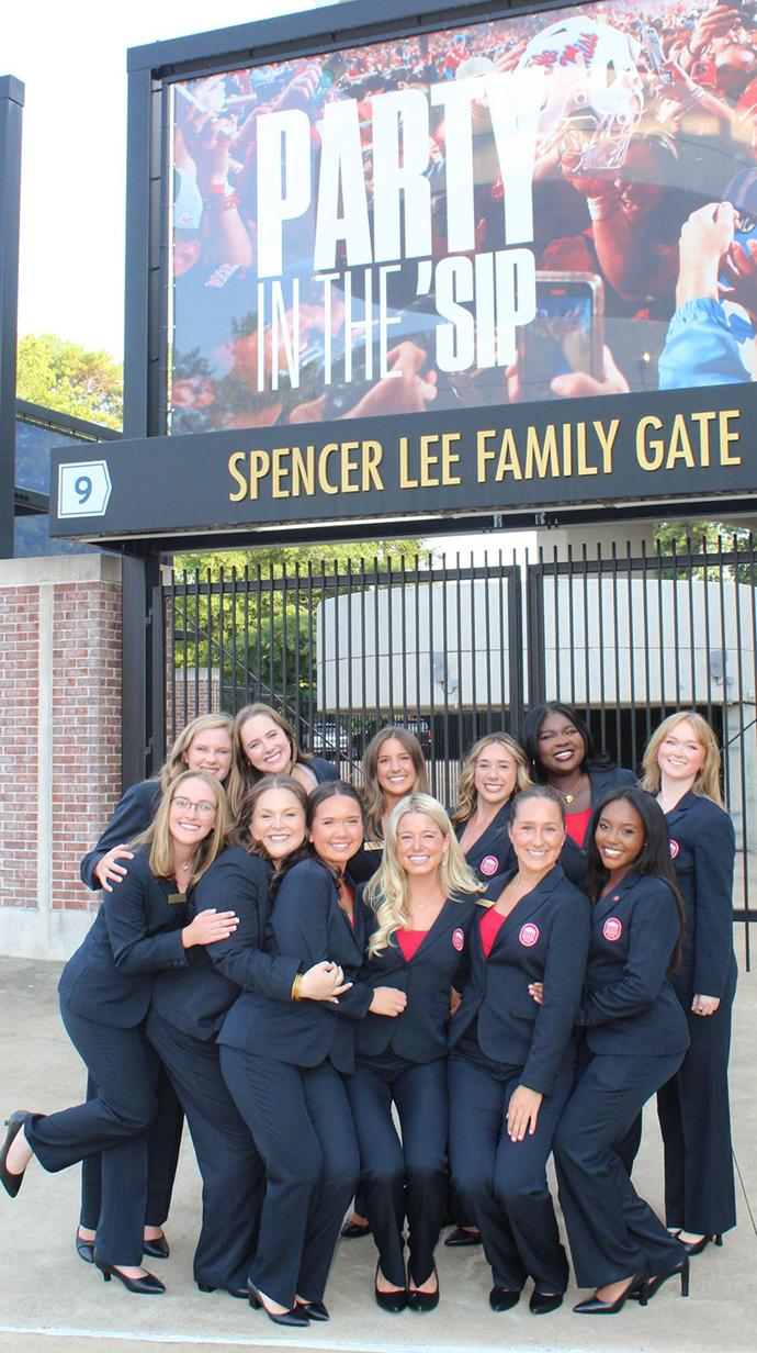 A group of young women, all wearing navy blue blazers, stand outside the entry gates of a stadium.