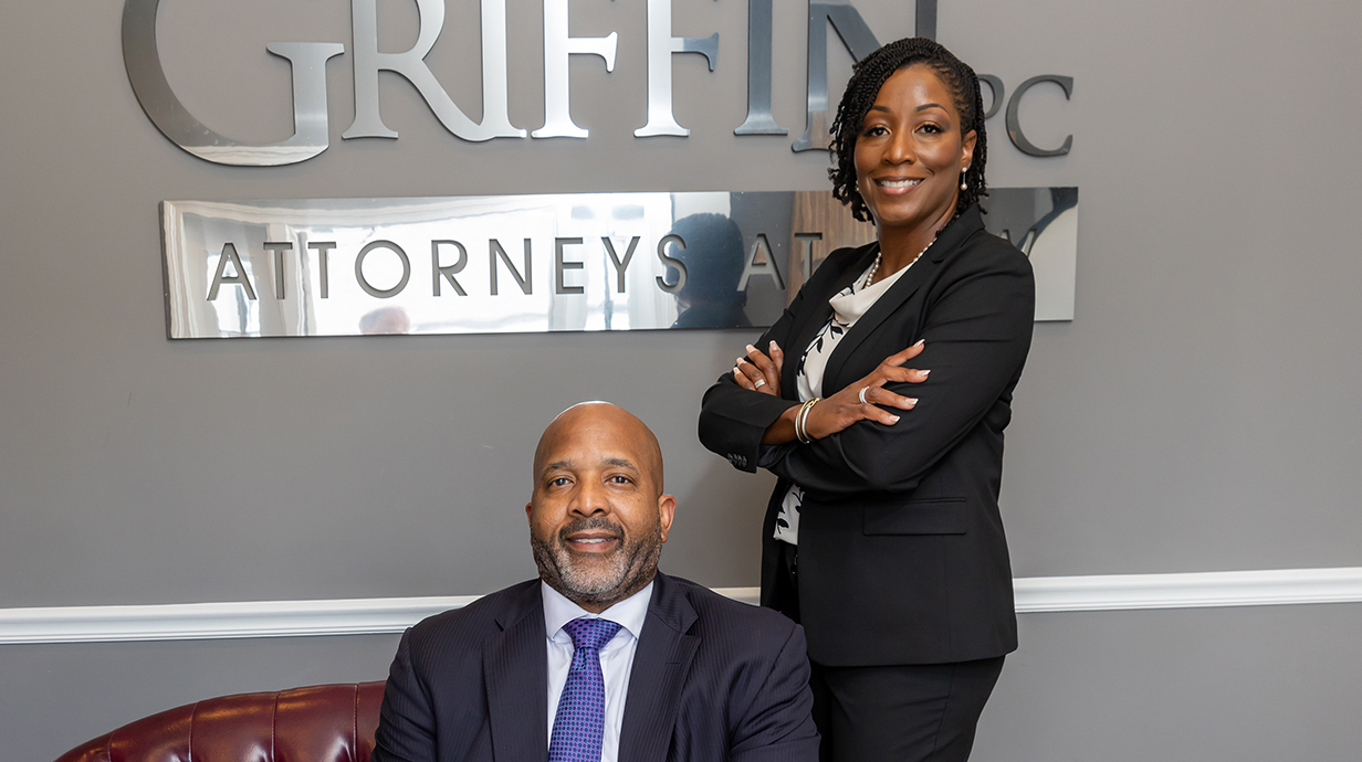 A woman stands as a man sits in front of a silver sign for a law firm on an earth-colored wall.