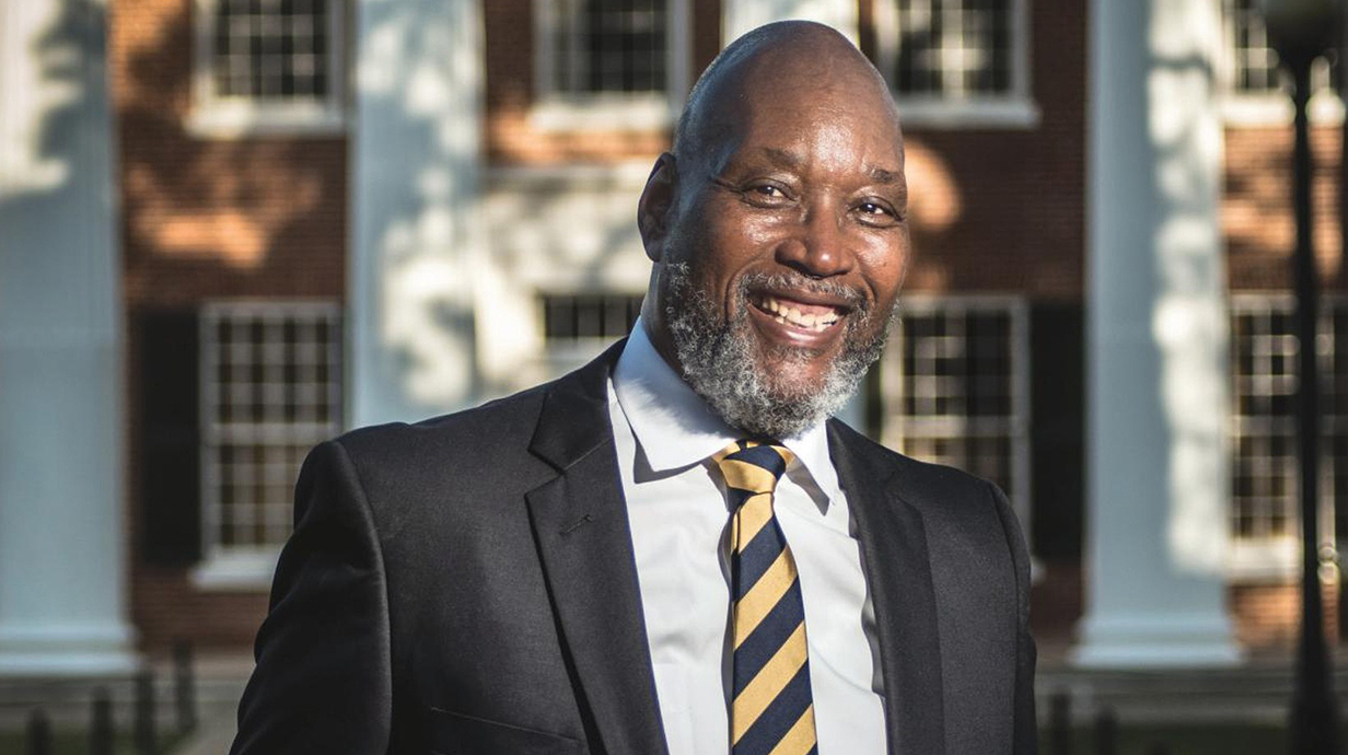 Photo of a man wearing a suit standing outside a large brick building with white columns.