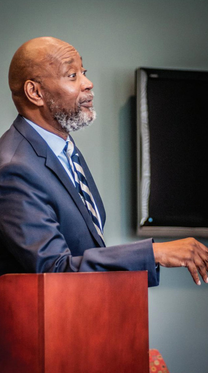 A man leans against a lecture as he talks to a classroom.
