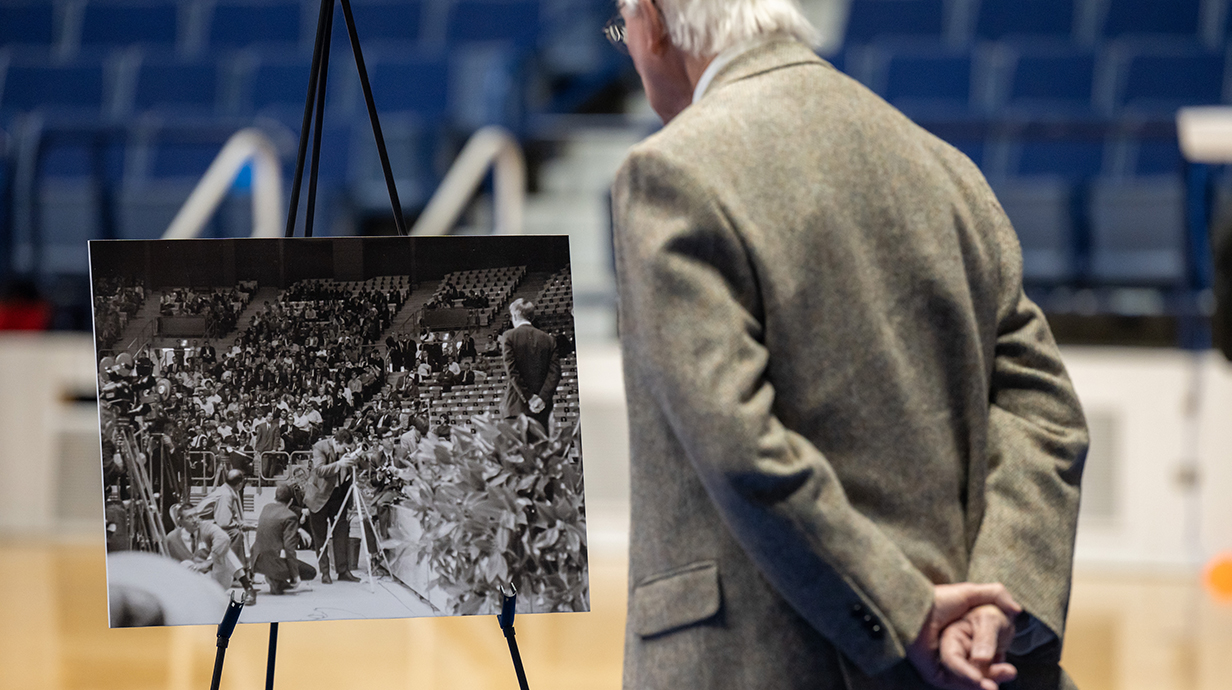 A man wearing a suit stands with his hands clasped behind his back as he views a black-and-white photograph in an arena.