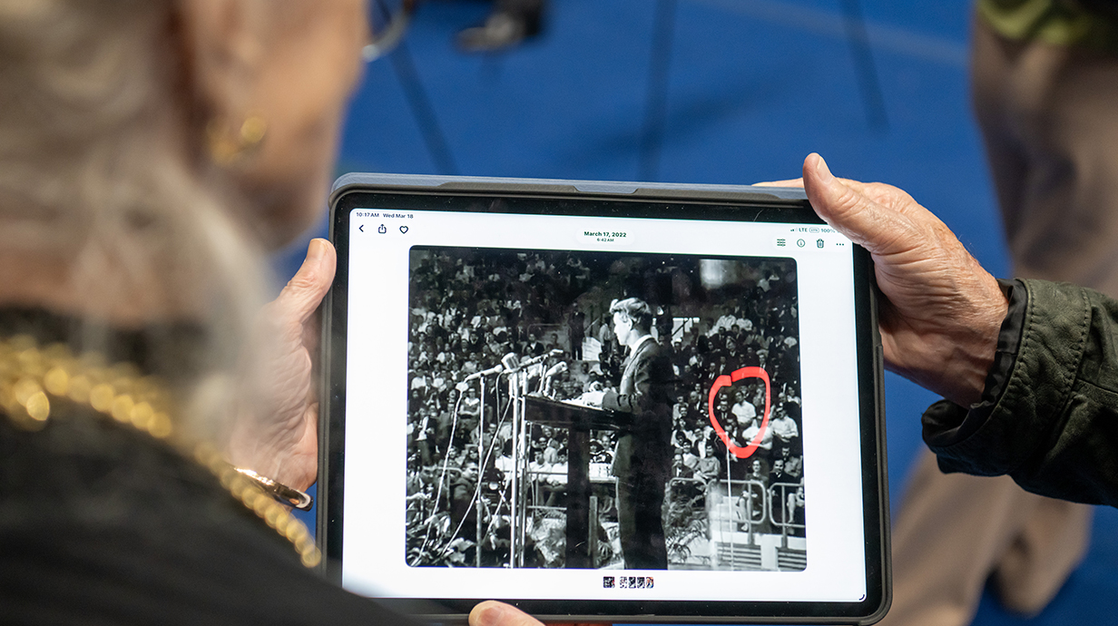 A hand points to a person circled in red on a photograph displayed on a tablet computer.