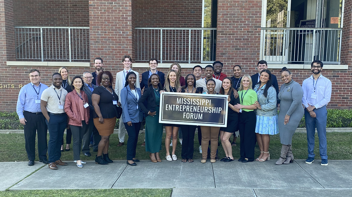 A group of people holding a banner that reads 'Mississippi Entrepreneurship Forum' stands in front of a large brick building.