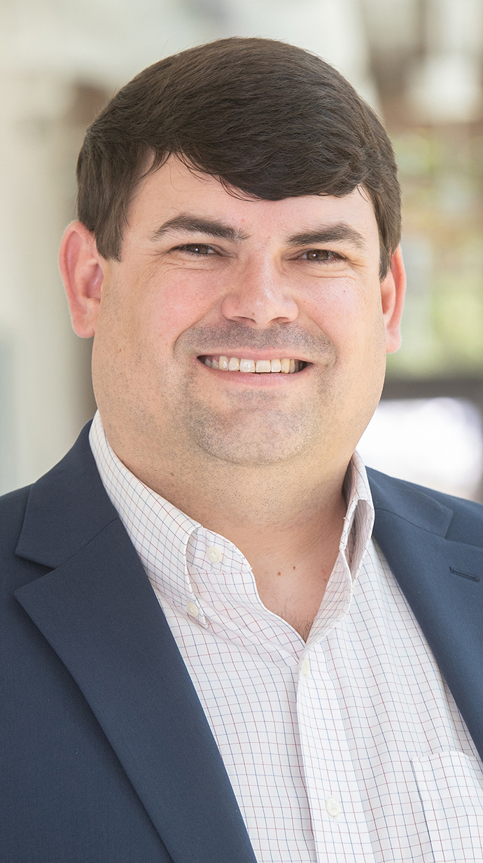 Headshot of a man wearing a dark blue jacket over a white shirt with blue checks.