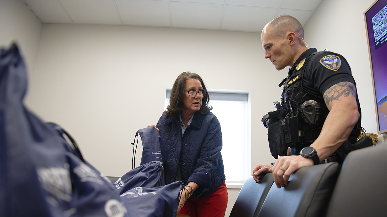 A woman shows a man dressed in a police uniform a pile of blue string backpacks on a table.