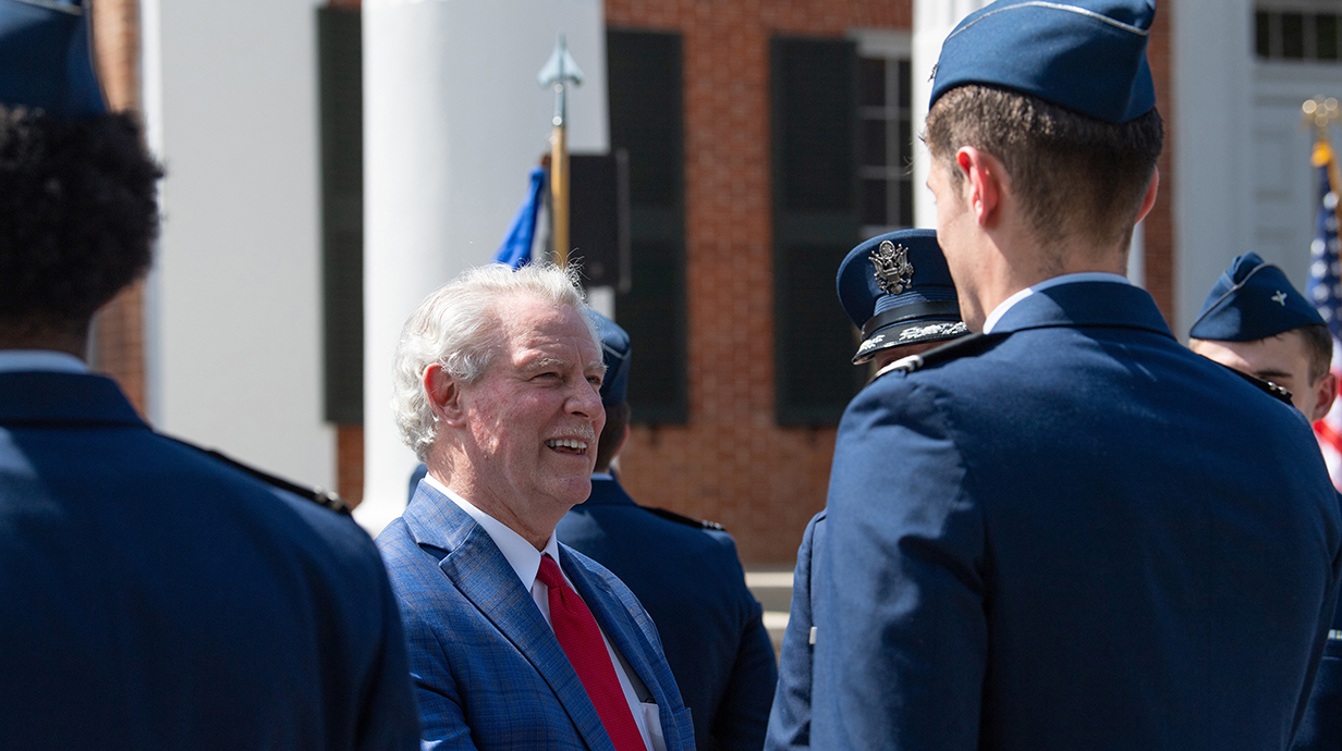 A man wearing a dark blue suit stands in front of several young men wearing military dress uniforms.