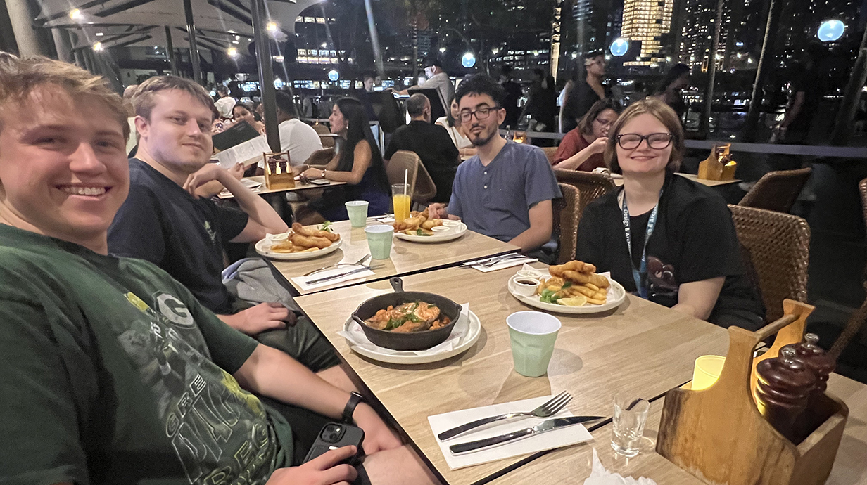 A roup of young people sit around a dining table with a city skyline behind them.