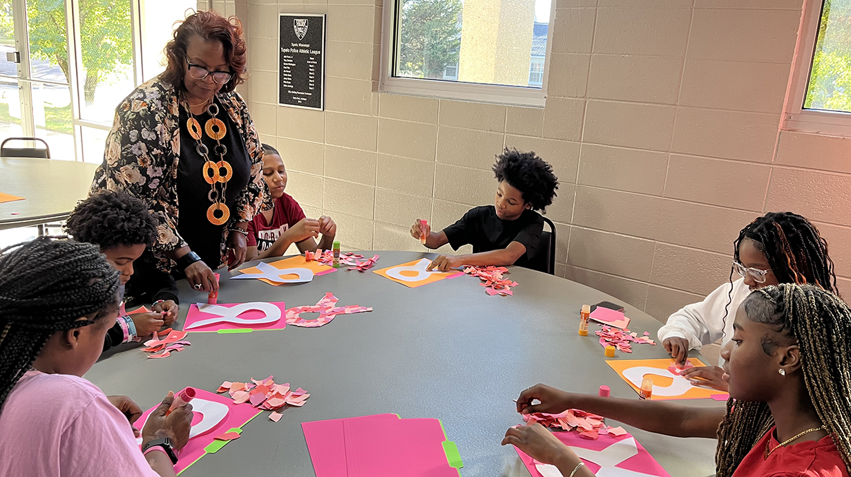 A woman standing in a classroom instructs a group of young people seated around a table on an art project.