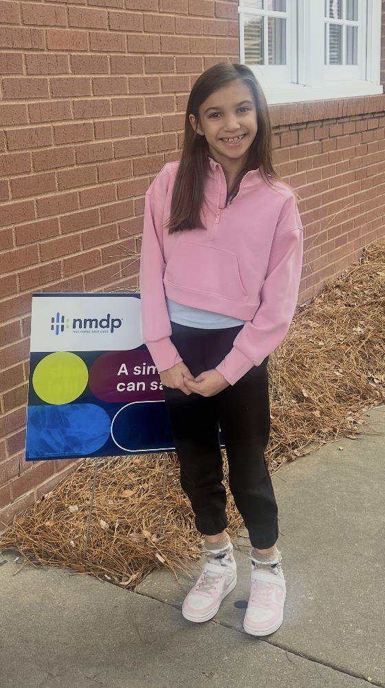 A young girl wearing a pink shirt stands outside a brick building.