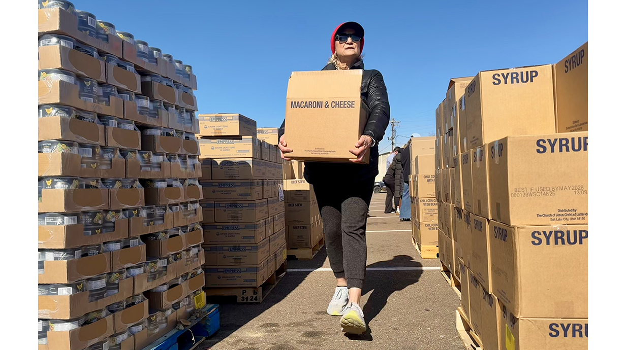 A woman wearing sunglasses carries a box of macaroni and cheese through a maze of food boxes stacked in a parking lot.