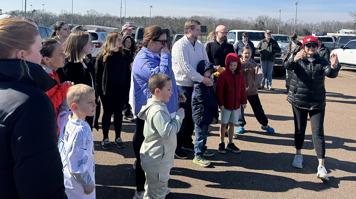 A woman gestures as she directs a group of people standing in a parking lot.