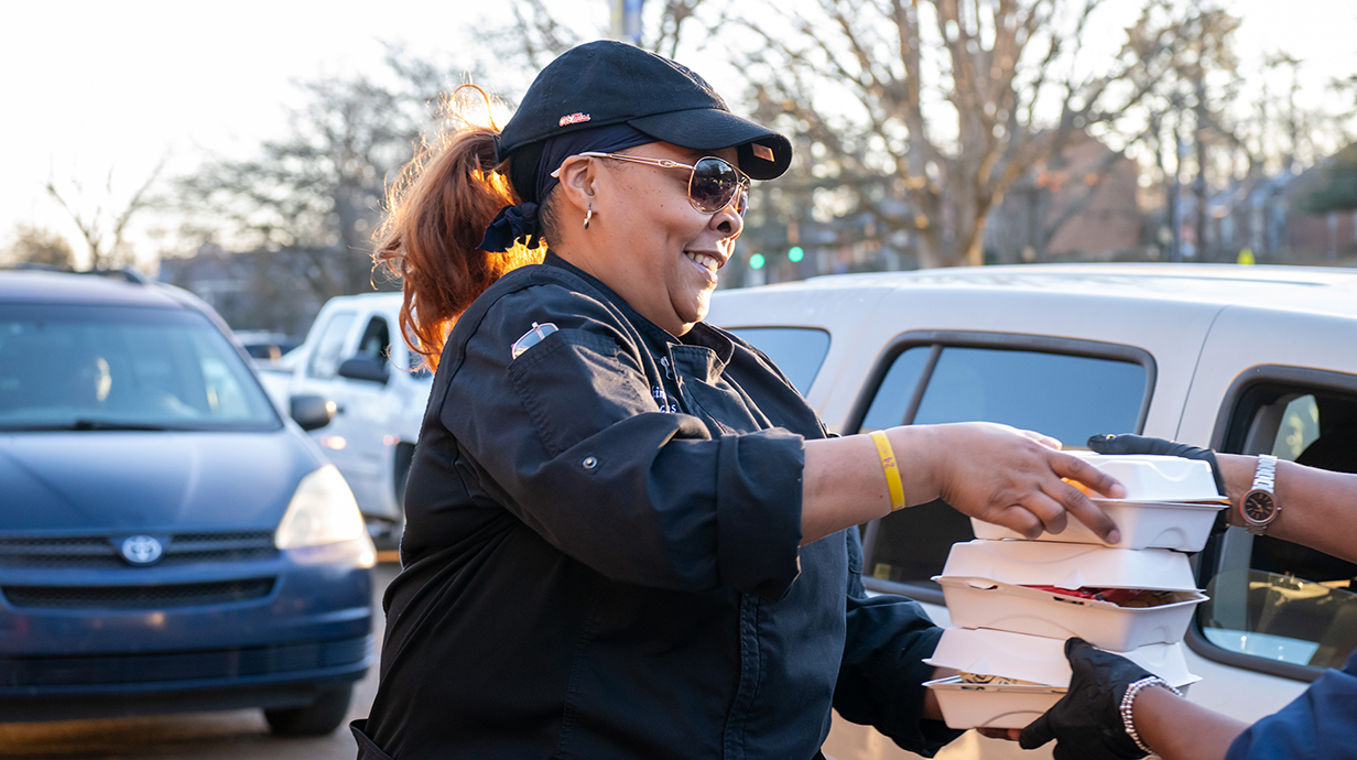 A woman wearing a baseball cap and a black chef's jacket hands out foam containers of food in a parking lot.