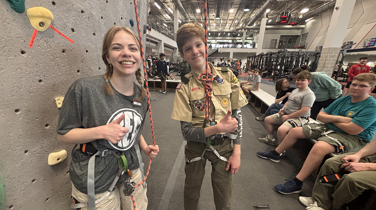 A young woman and a young man wearing a Scout uniform give a thumbs-up while wearing harnesses and standing next to a climbing wall.