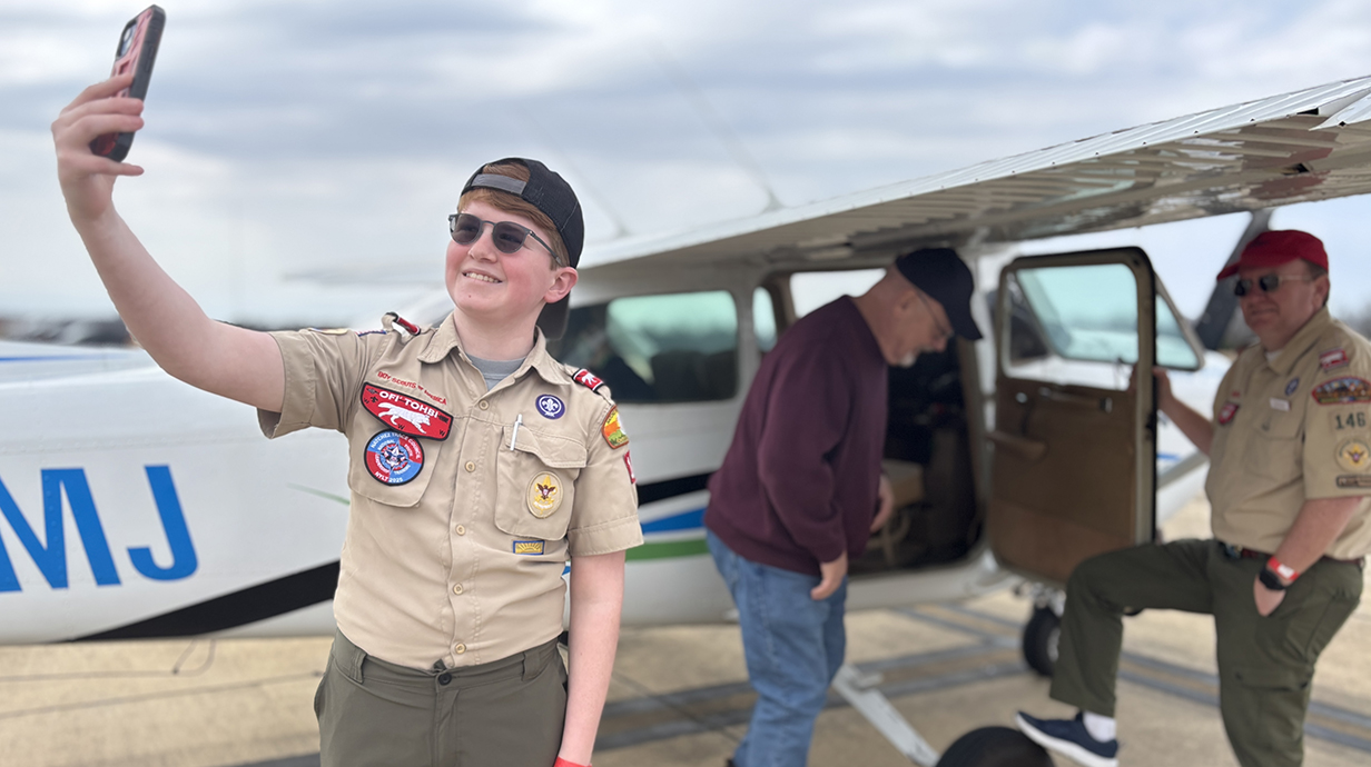 A young man in a Scout uniform takes a selfie in front of an airplane with two men standing nearby.