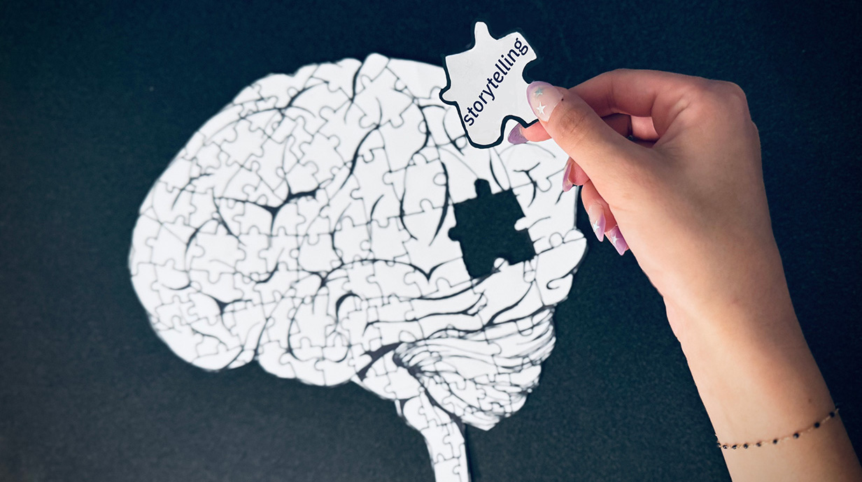 Photo of a hand holding a jigsaw puzzle piece with the word 'Storytelling' written on it above a nearly completed image of a human brain.